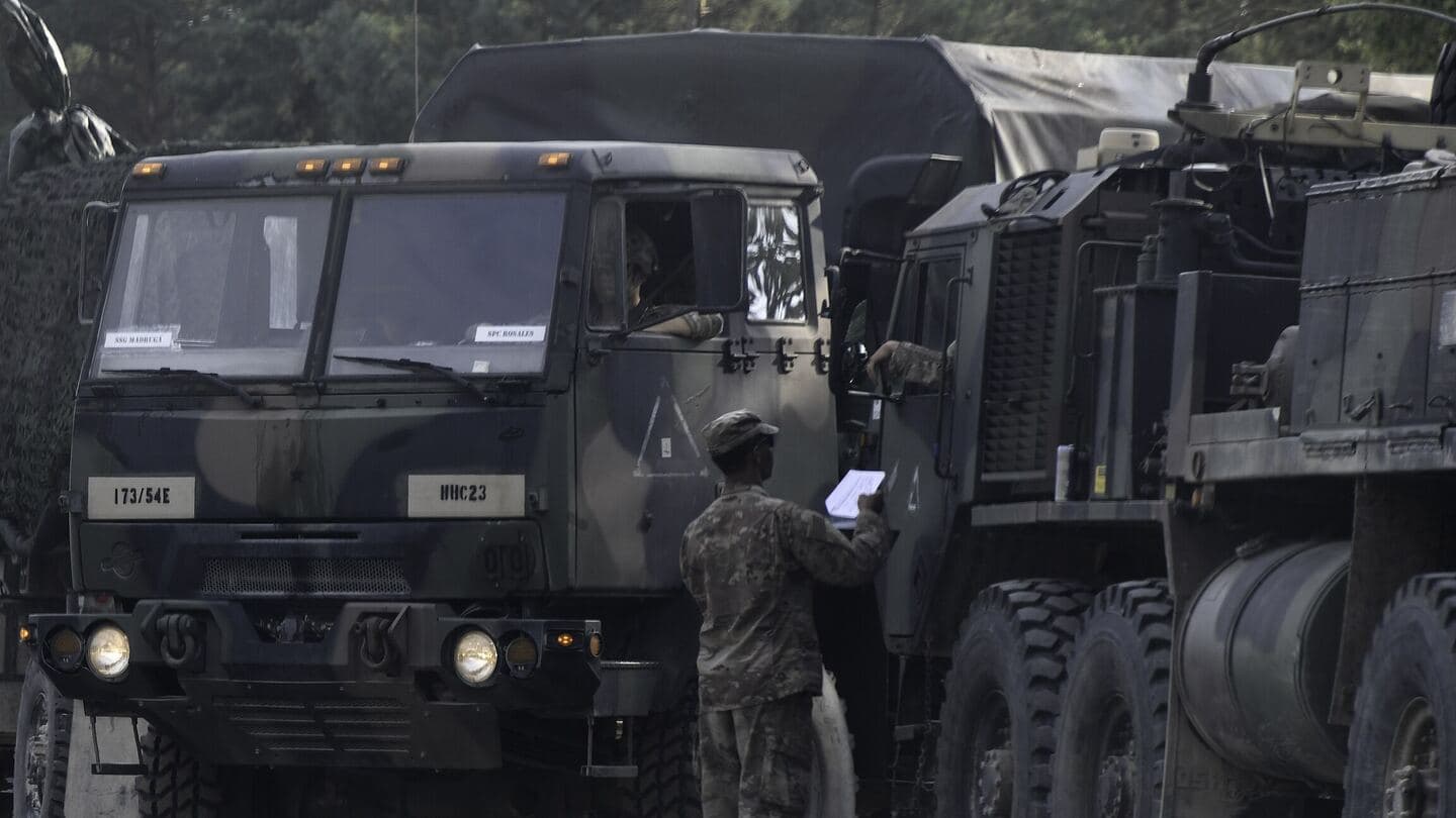 A soldier checks paperwork at a line of camouflaged Army trucks staged for a convoy