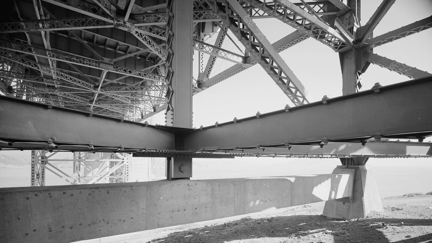 Steel trestle trusses beneath a suspension bridge approach, seen from below in black and white