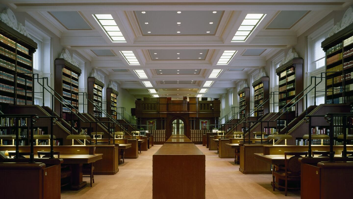 A long library reading room with desks, reading lamps and shelved collections under a coffered ceiling