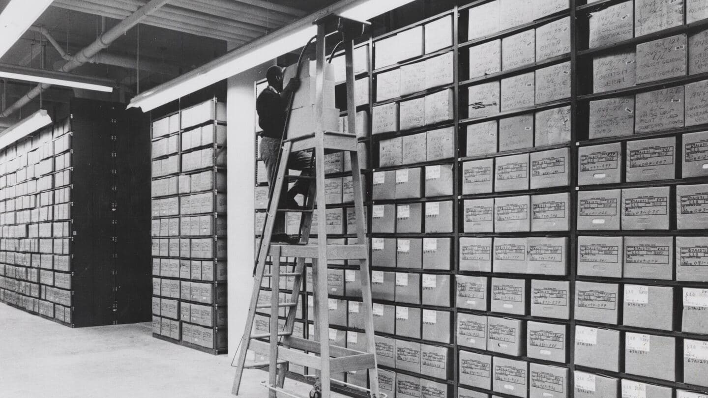 A records center stack area with floor-to-ceiling shelves of boxed files and a worker on a ladder