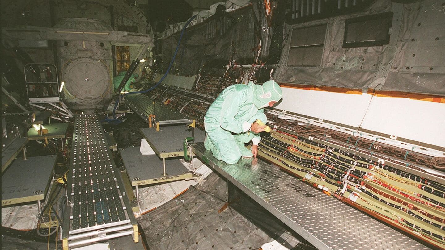 A technician in a clean-room suit inspecting wiring bundles along an orbiter payload bay