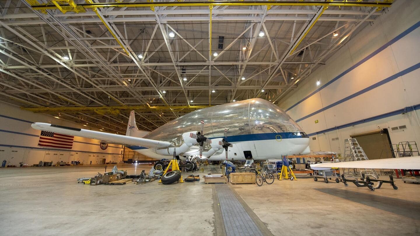 A large cargo aircraft on jacks in a hangar, with a wheel removed and parts laid out for maintenance