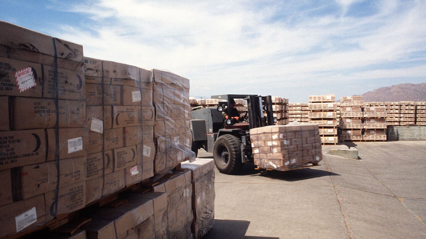 A forklift moves a pallet of boxed supplies past rows of stacked pallets in an outdoor storage yard