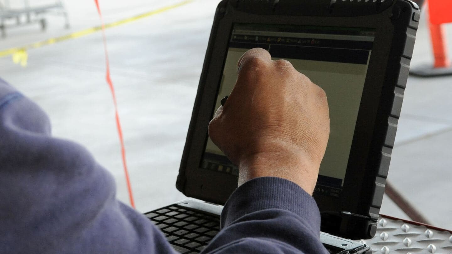 A hand uses a stylus on a rugged convertible laptop screen beside an aircraft
