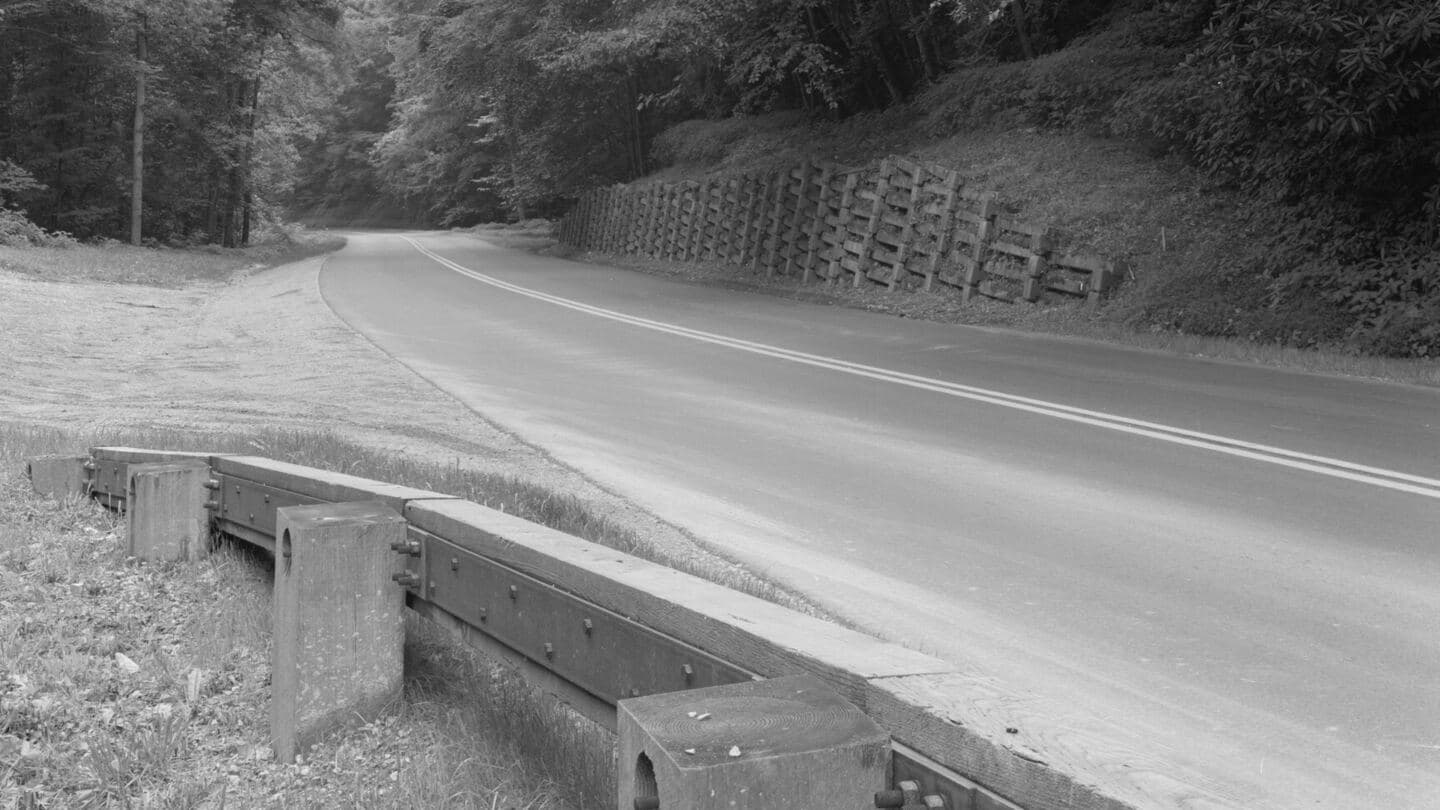 A steel-backed timber guardrail lining a curving mountain road, black and white