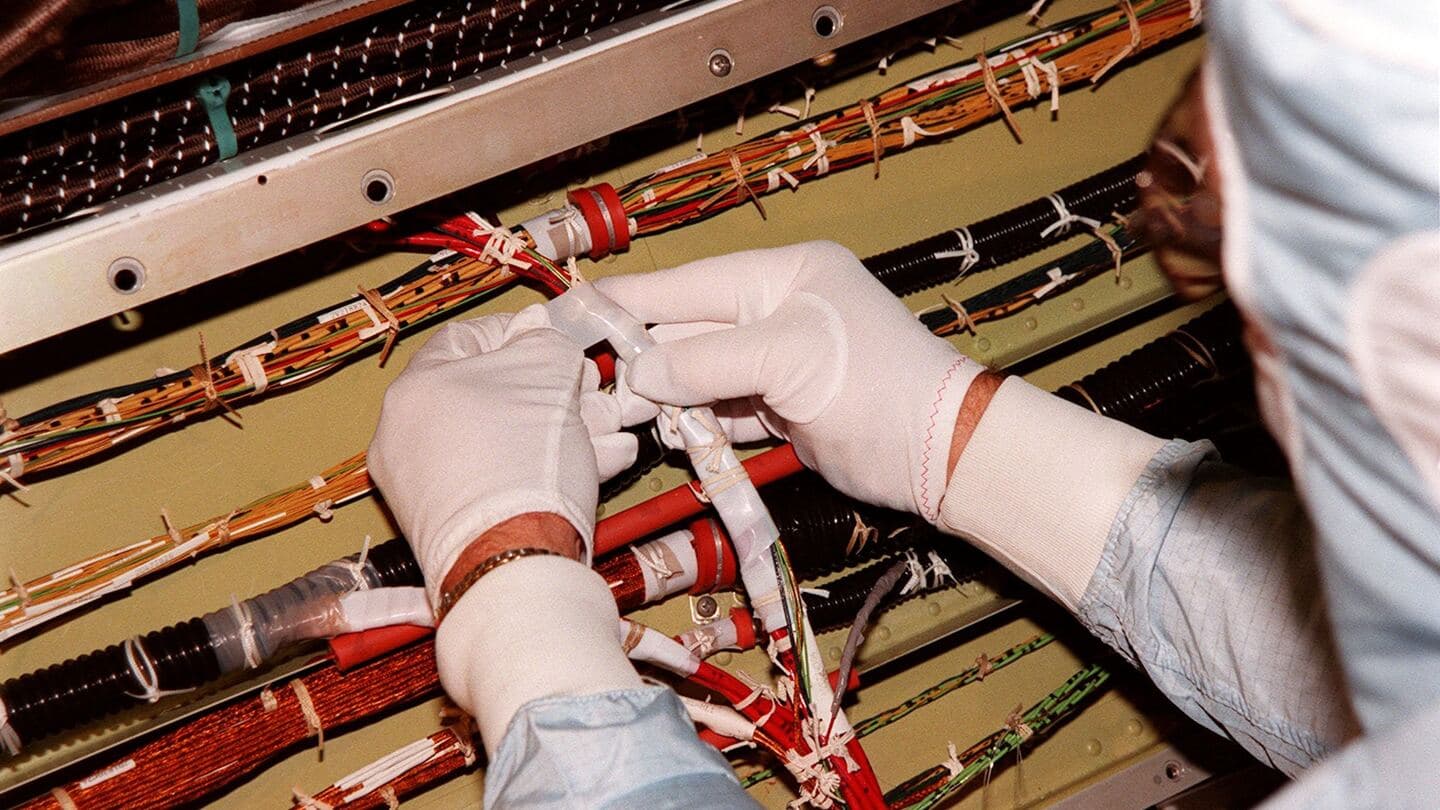 Gloved hands tracing a bundle of wires inside a spacecraft equipment bay during an inspection