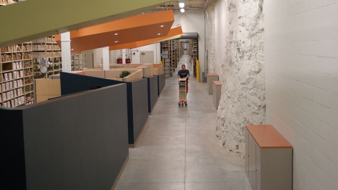 A records clerk pushes a cart down an aisle of shelved files in an underground records center