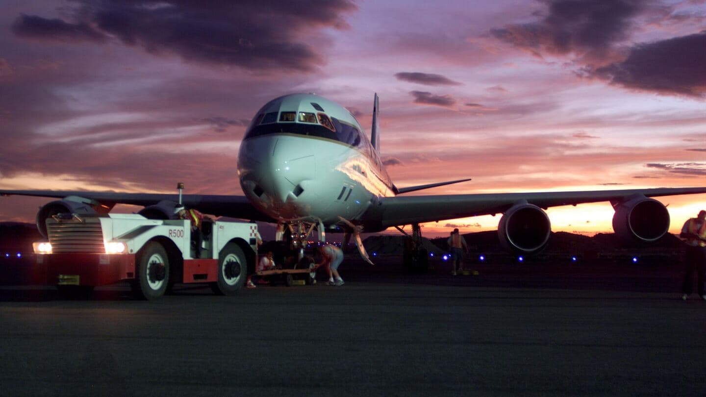 A four-engine jet on the ramp at sunset with a ground tug